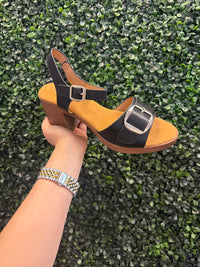 Hand holding a sandal with a buckle design against a green textured background