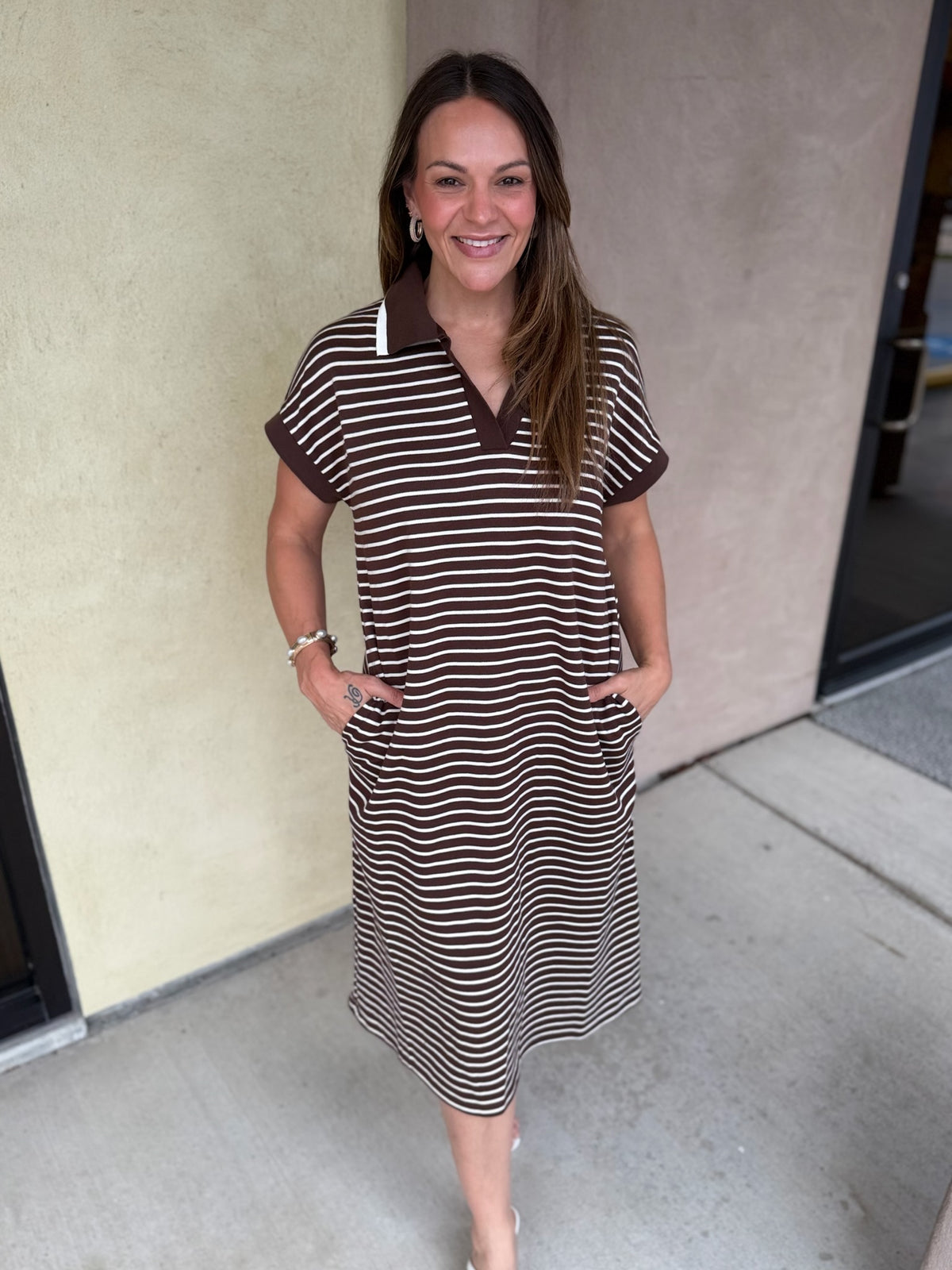 A woman modeling a brown and beige horizontally striped short-sleeve polo dress with a solid brown collar, standing outdoors against a beige stucco wall.