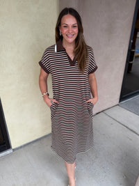 A woman modeling a brown and beige horizontally striped short-sleeve polo dress with a solid brown collar, standing outdoors against a beige stucco wall.