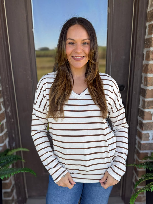 A woman smiling and wearing a cream-colored long-sleeve V-neck top with thin brown horizontal stripes, paired with blue jeans, standing in front of a rustic brick and wood door.