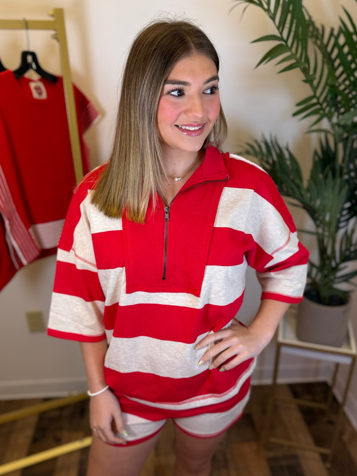 A woman wearing a red and off-white horizontally striped short-sleeve quarter-zip pullover top with matching shorts, standing in an indoor room with a plant and clothing rack visible in the background.
