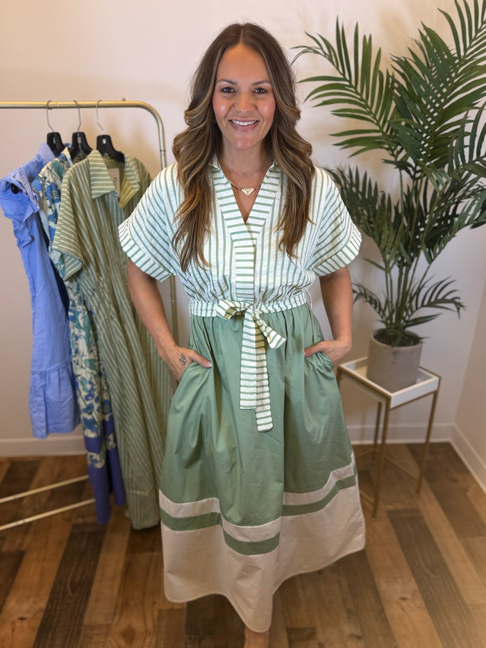 Woman wearing a sage and white midi dress with striped top and solid skirt standing in a room with clothing racks and plants.