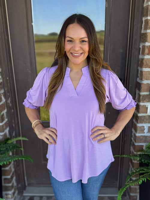 A woman wearing a lavender blouse with short puff sleeves, smocked detailing, and a split V neckline.