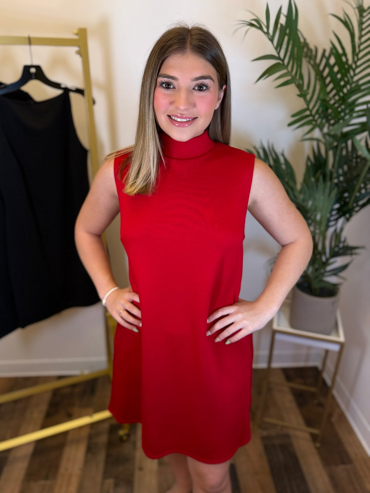A woman wearing a sleeveless, high-neck red dress stands indoors with her hands on her hips. A black dress is hanging on a gold clothing rack to the left, and a potted plant is on a small table to the right.