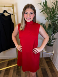 A woman wearing a sleeveless, high-neck red dress stands indoors with her hands on her hips. A black dress is hanging on a gold clothing rack to the left, and a potted plant is on a small table to the right.