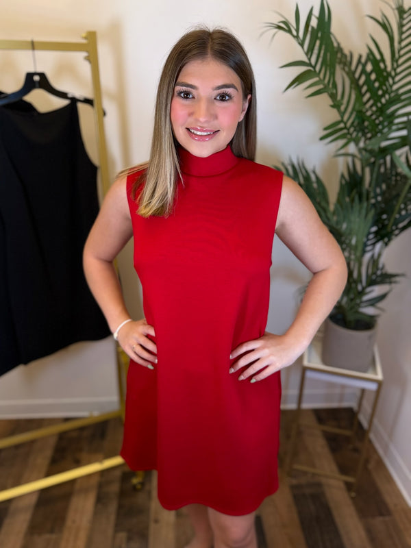 A woman wearing a sleeveless, high-neck red dress stands indoors with her hands on her hips. A black dress is hanging on a gold clothing rack to the left, and a potted plant is on a small table to the right.