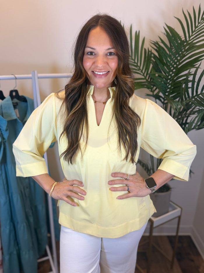 Woman wearing a pale yellow blouse with quarter length sleeves and ruffled neckline in a clothing store setting.