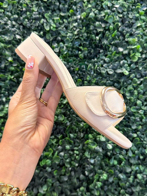 Beige high-heeled shoe with decorative gold element held against a green leafy background