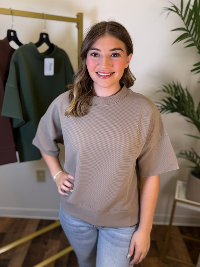 A woman wearing a taupe short-sleeve crewneck sweatshirt and light blue jeans, standing in a boutique setting with a clothing rack and plant in the background.