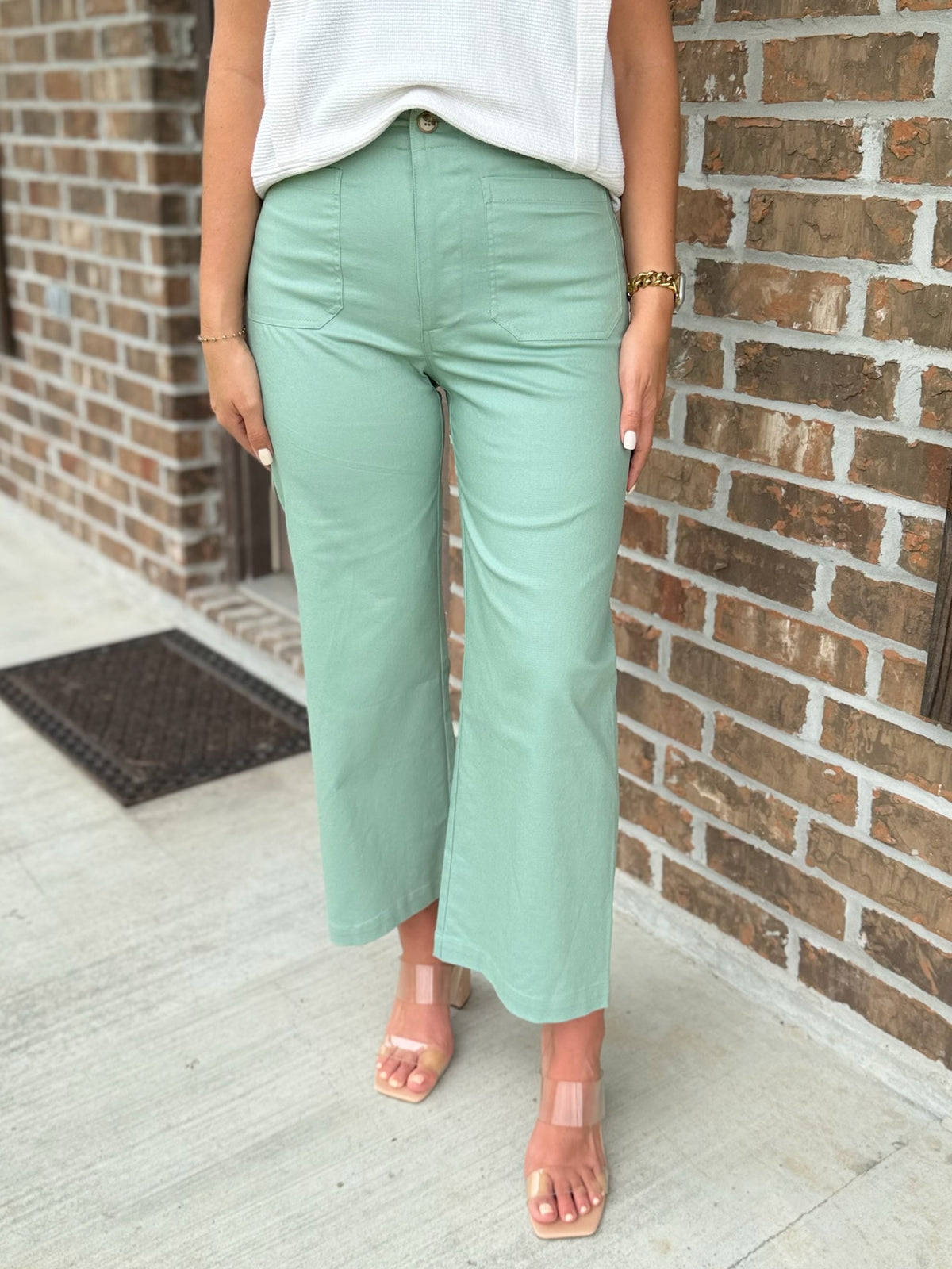 Person wearing light green wide leg pants and a white top against a brick wall.