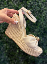 Beige wedge sandals with espadrille sole held against a green leafy background