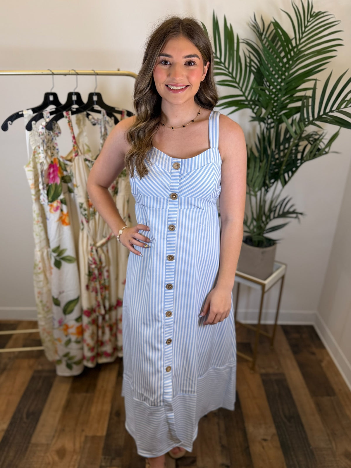Woman wearing a sleeveless blue striped dress with button front details in a room with clothing racks and a plant.