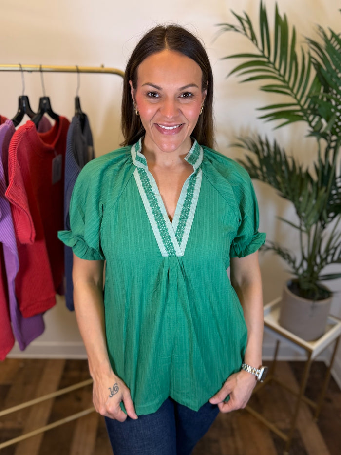 A woman wearing a green short, puff sleeve blouse with white embroidered trim on the V-neckline, standing in a boutique setting with clothing racks and a potted plant in the background.