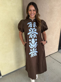 A woman wearing a brown midi dress with puff sleeves, a mock neck, and a light blue vertical damask pattern on the front, standing against a beige stucco wall.