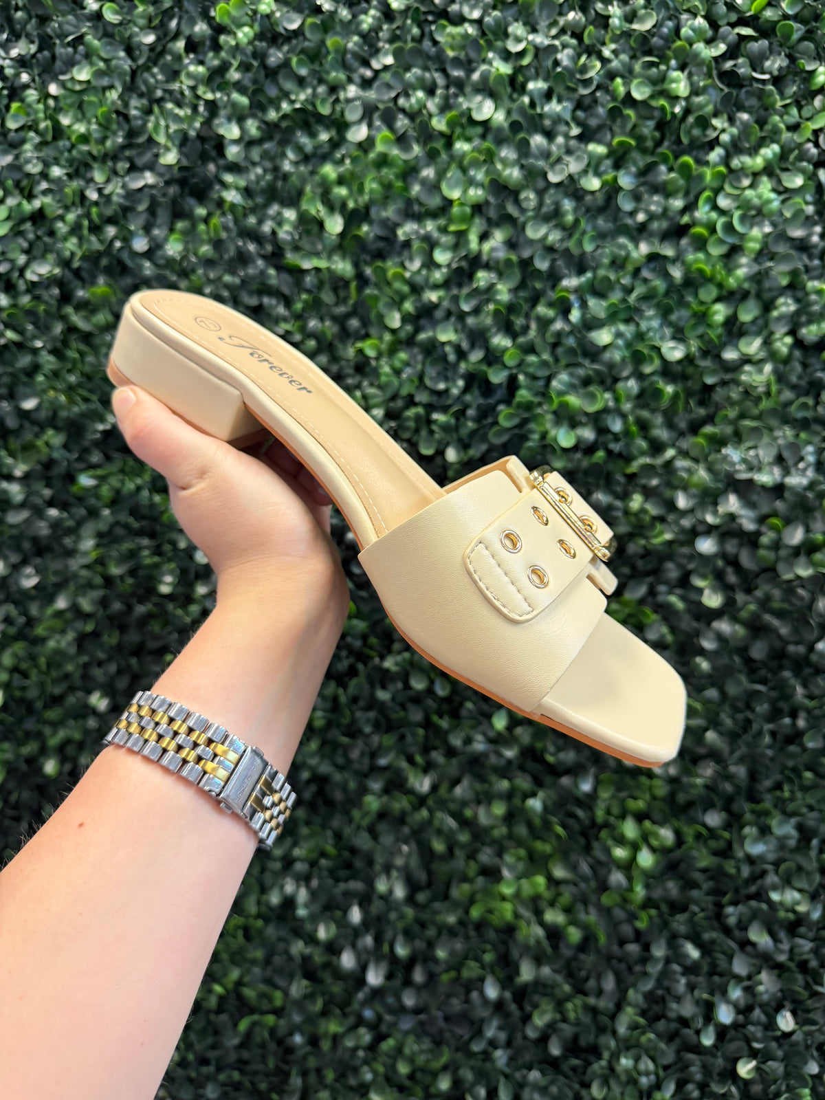 Beige slip on sandal with short block heel held by a hand against a green textured background