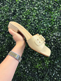 Beige slip on sandal with short block heel held by a hand against a green textured background
