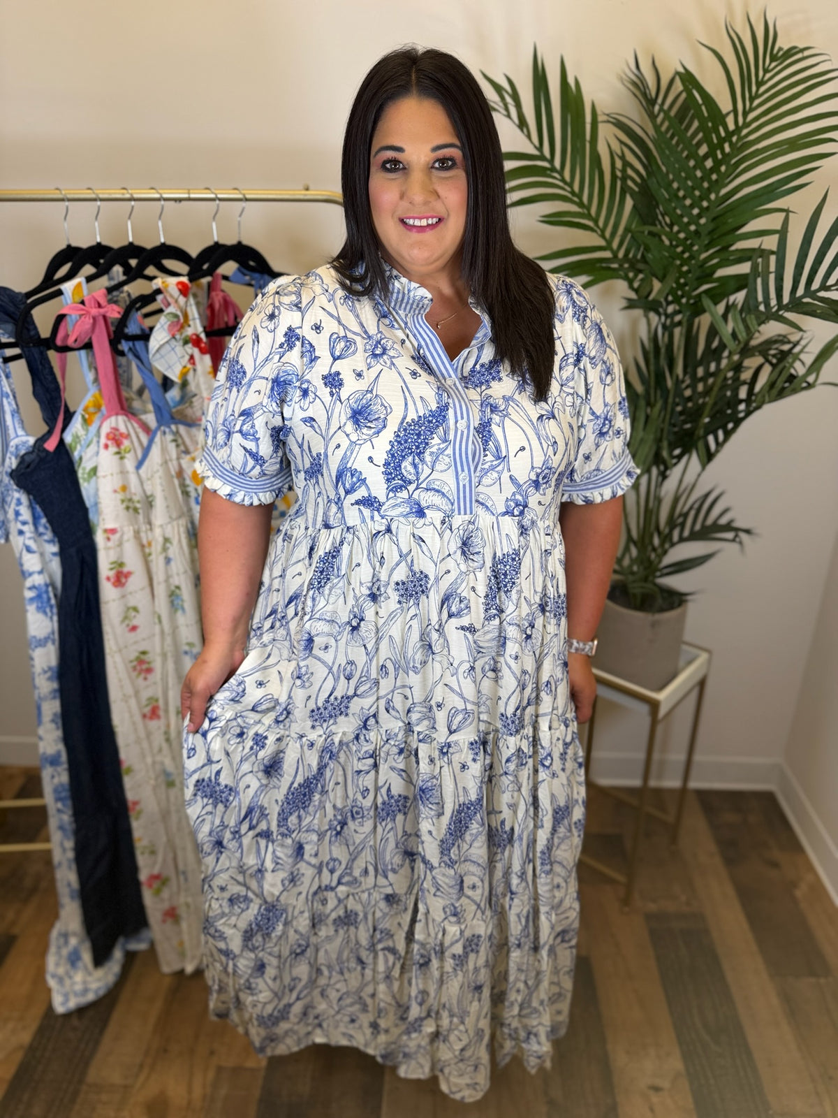 Woman wearing a blue and white floral maxi dress with striped contrast trim and button details standing in a room with clothes on hangers and a plant.