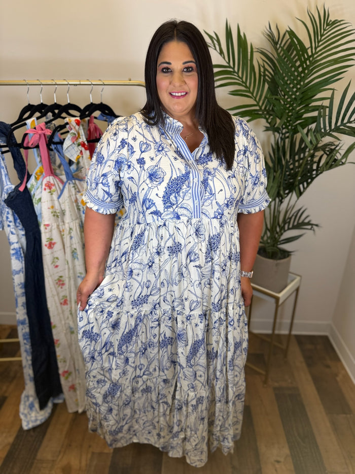 Woman wearing a blue and white floral maxi dress with striped contrast trim and button details standing in a room with clothes on hangers and a plant.