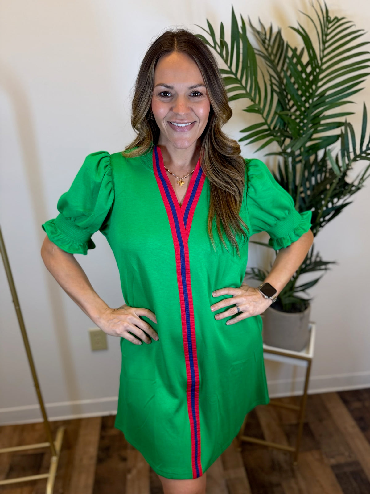 Woman wearing a puff sleeve green dress with red and blue accents, standing indoors with a plant in the background.