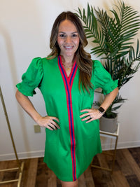 Woman wearing a puff sleeve green dress with red and blue accents, standing indoors with a plant in the background.
