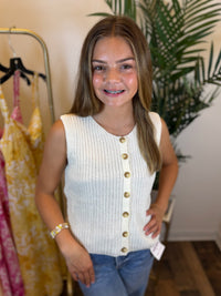 Young girl wearing a white sleeveless button-up knit top with tan buttons in a clothing store.