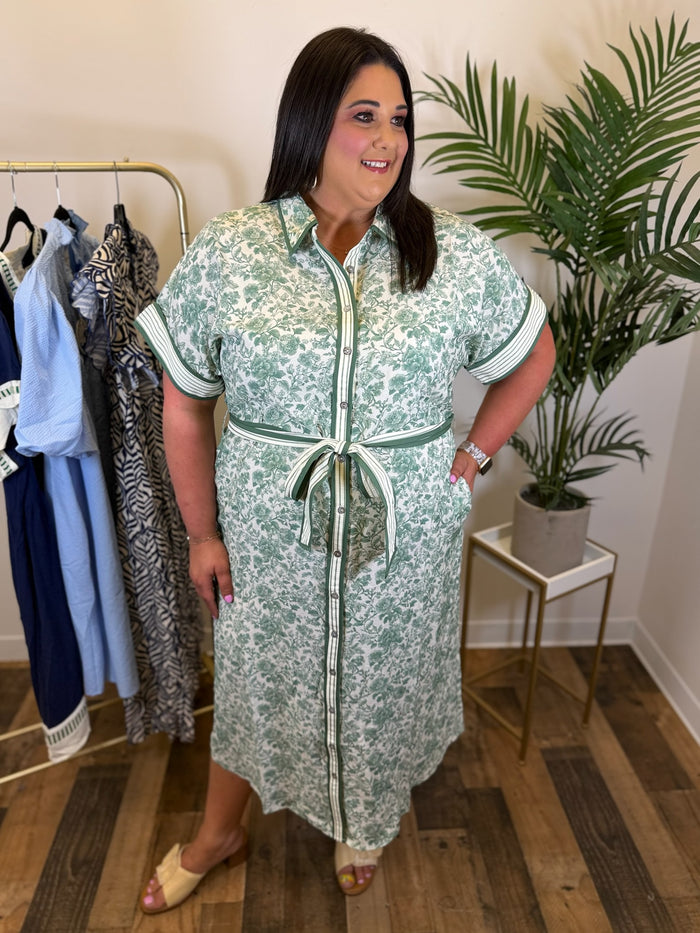 Woman wearing a green floral button front dress with contrast trim details standing in a room with clothes and plants.
