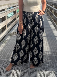 Person wearing navy patterned pants with a wide, flowy fit and elastic waistband standing on a wooden deck.