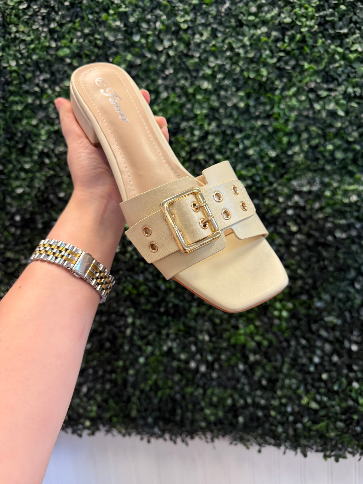 Beige slip on heel with a gold buckle held by a hand against a green bush background