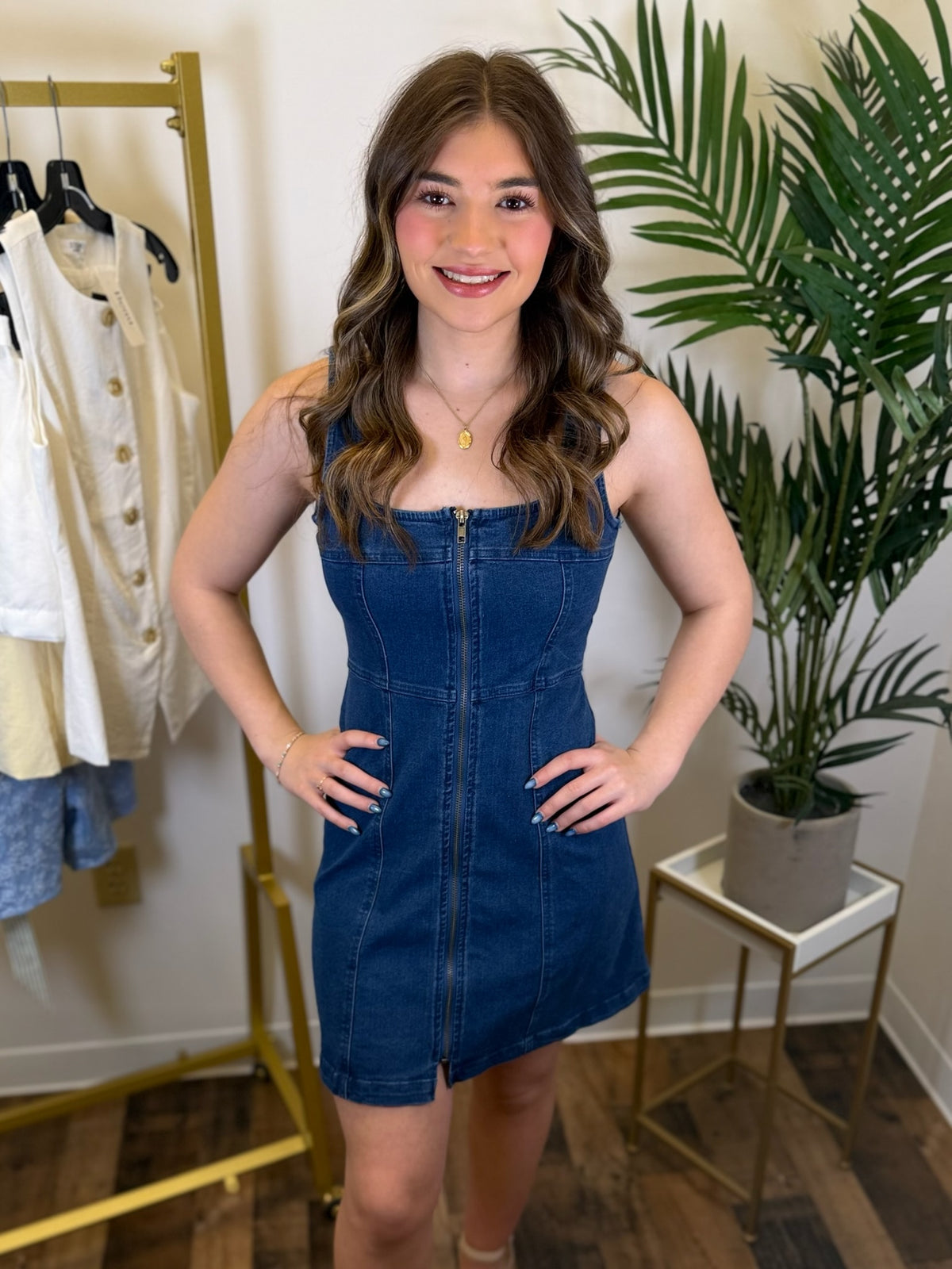 Woman wearing a blue denim dress standing in a room with a clothing rack and plant.