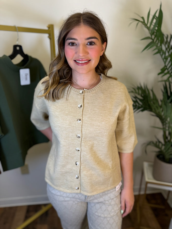 A woman modeling a short-sleeve knit cardigan in a light beige color, featuring a decorative button-down front. She is standing in a boutique setting with a clothing rack and a potted plant visible in the background.