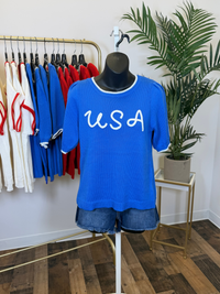 Mannequin wearing a blue 'USA' t-shirt with a plant and clothing rack in the background.