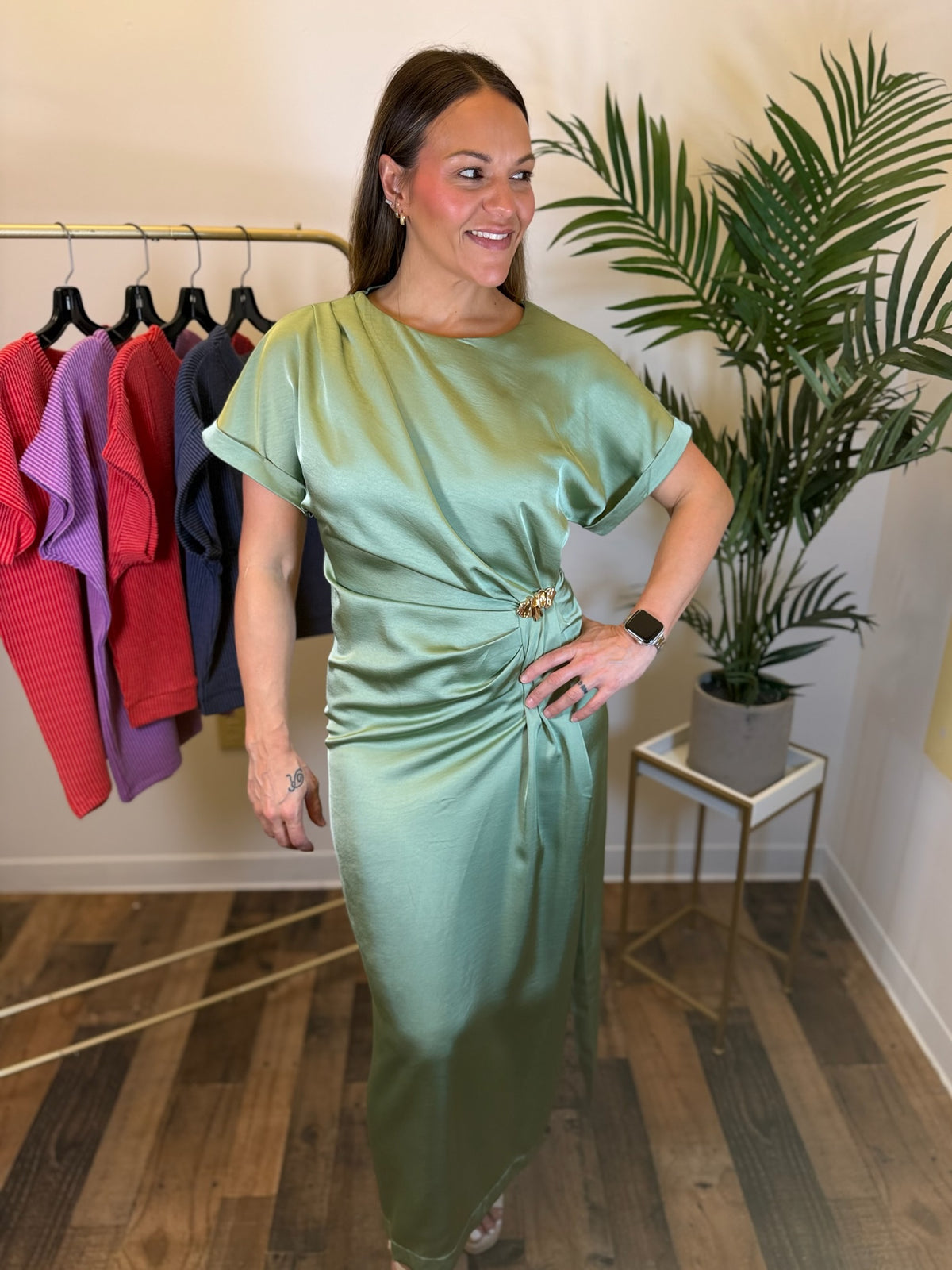 A woman modeling a sage green satin midi dress with short sleeves and a gathered waist detail, standing in a boutique setting with a clothing rack and a potted plant in the background.
