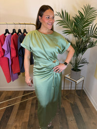 A woman modeling a sage green satin midi dress with short sleeves and a gathered waist detail, standing in a boutique setting with a clothing rack and a potted plant in the background.
