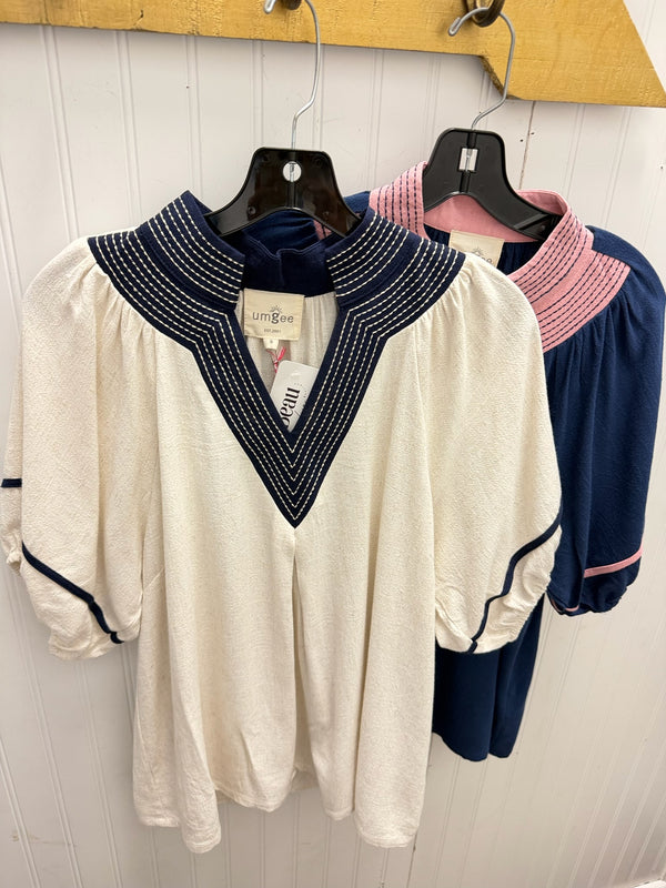 An off-white short-sleeve blouse with a navy blue V-neckline and sleeve trim, hanging on a black hanger against a white paneled wall. A second blouse in navy and pink is partially visible behind it.