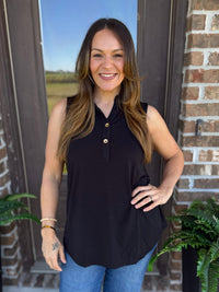 A woman wearing a sleeveless black polo-style top with gold buttons and blue jeans, standing in front of a brick doorway with plants visible on both sides.