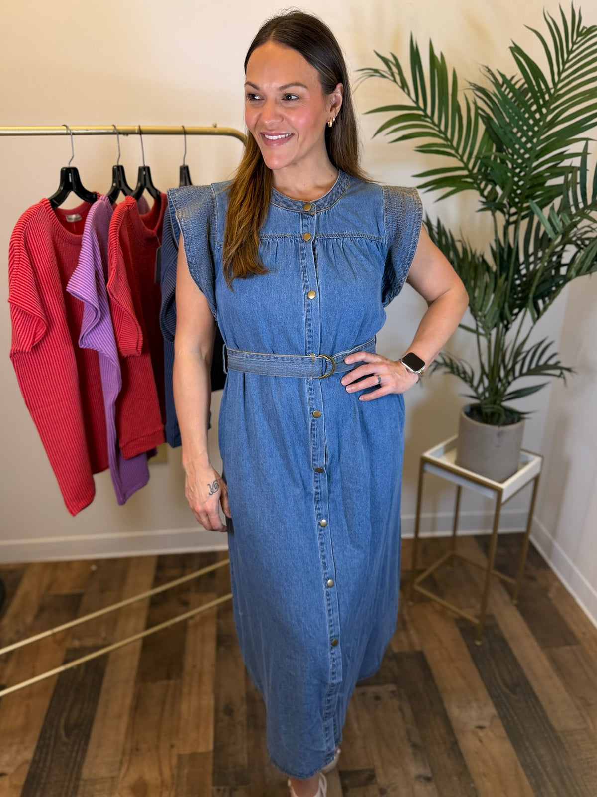 A woman modeling a sleeveless blue denim midi dress with a button-down front and matching belt, standing in a boutique setting with a clothing rack and potted plant in the background.