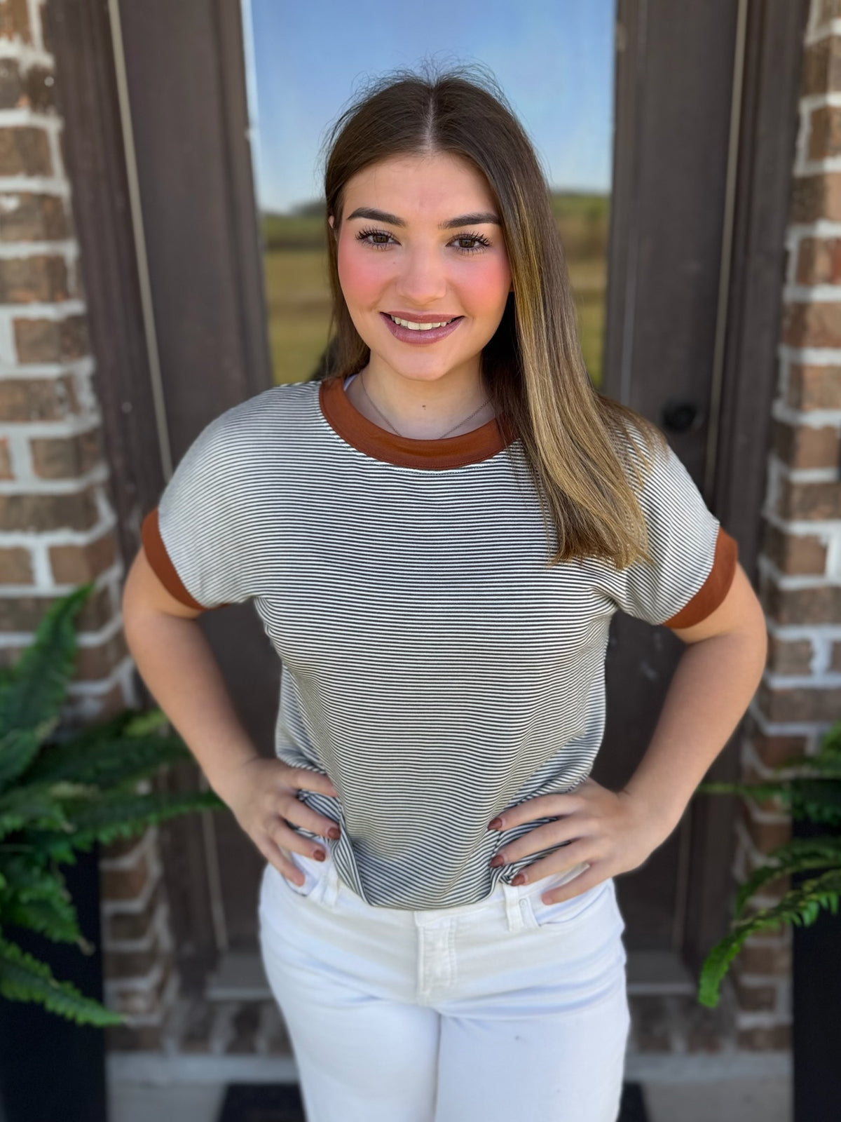 A woman wearing a short-sleeve ringer t-shirt with thin olive and white horizontal stripes and brown collar and sleeve trim, paired with white pants, standing in front of a brick door.