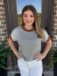 A woman wearing a short-sleeve ringer t-shirt with thin olive and white horizontal stripes and brown collar and sleeve trim, paired with white pants, standing in front of a brick door.