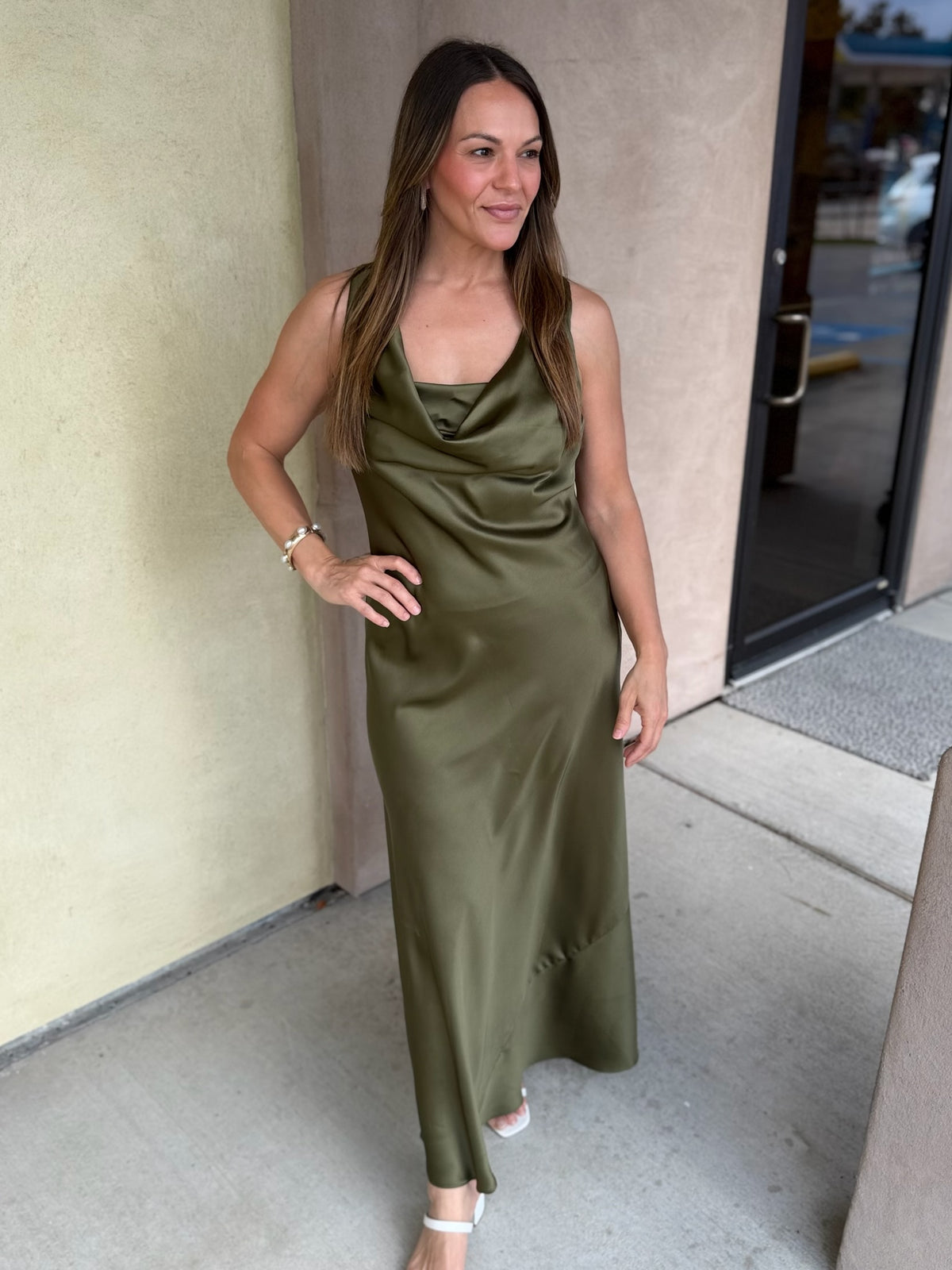 Olive green satin midi dress with spaghetti straps and draped neckline worn by a woman standing in front of beige building.