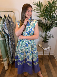 Woman wearing a floral midi dress standing in a room with a rack of clothes and a plant.
