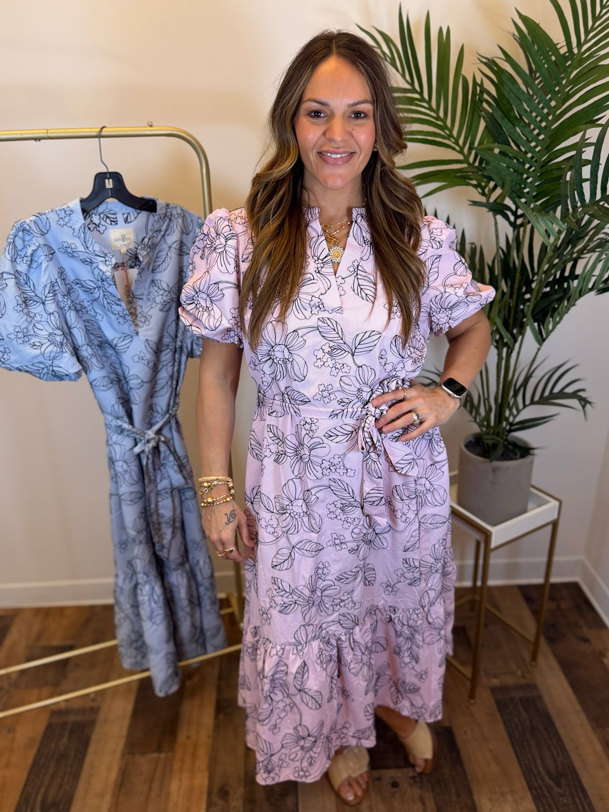 Woman wearing a pink floral dress midi dress with split neckline and short sleeves standing next to a blue robe in a room with a plant.