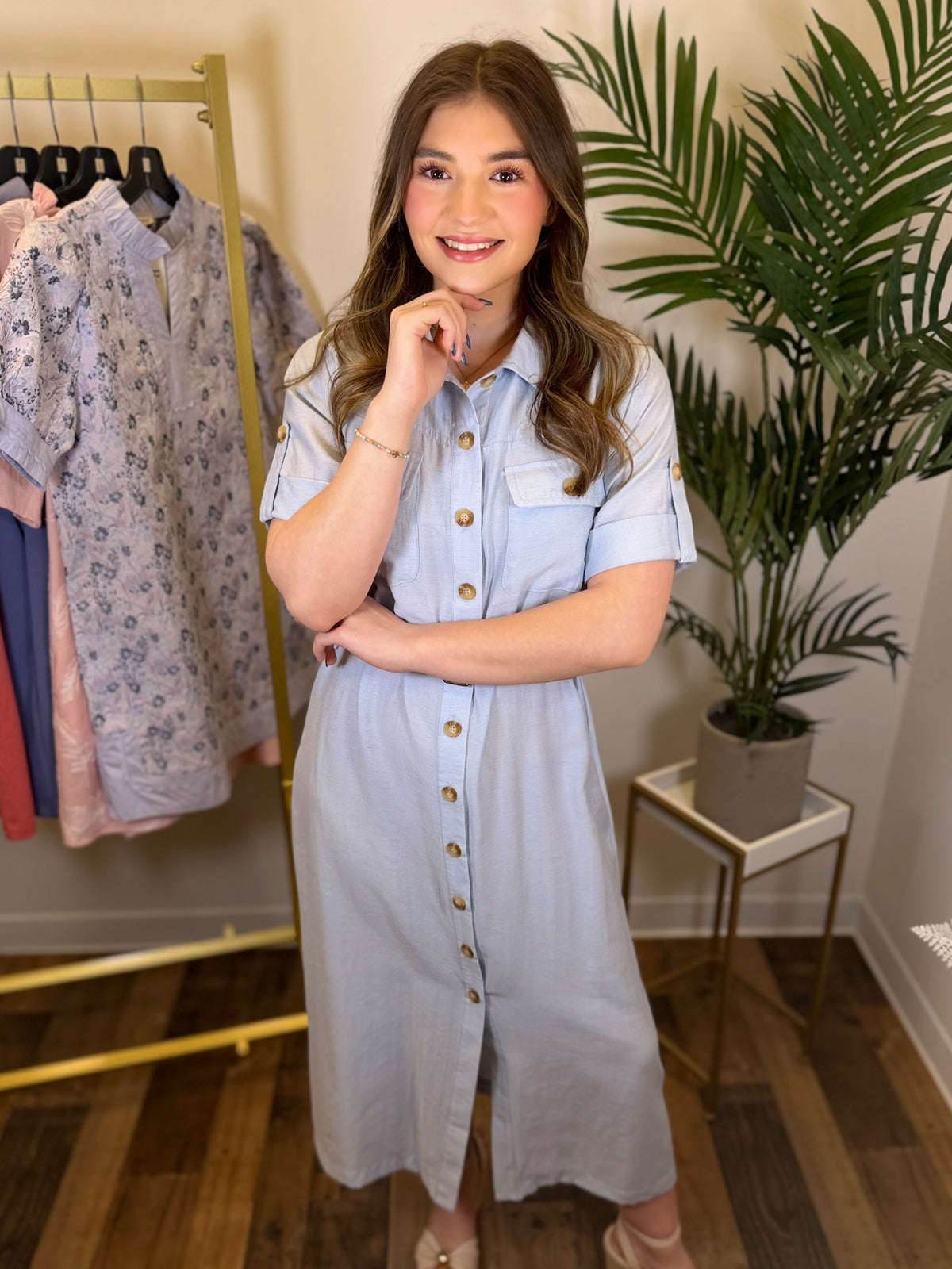 Woman in a light blue dress standing in a room with a rack of clothes and a plant.