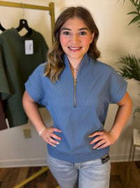 A woman modeling a dusty blue short-sleeve top with a half-zip mock neck, standing in a boutique setting with a clothing rack and plant in the background.