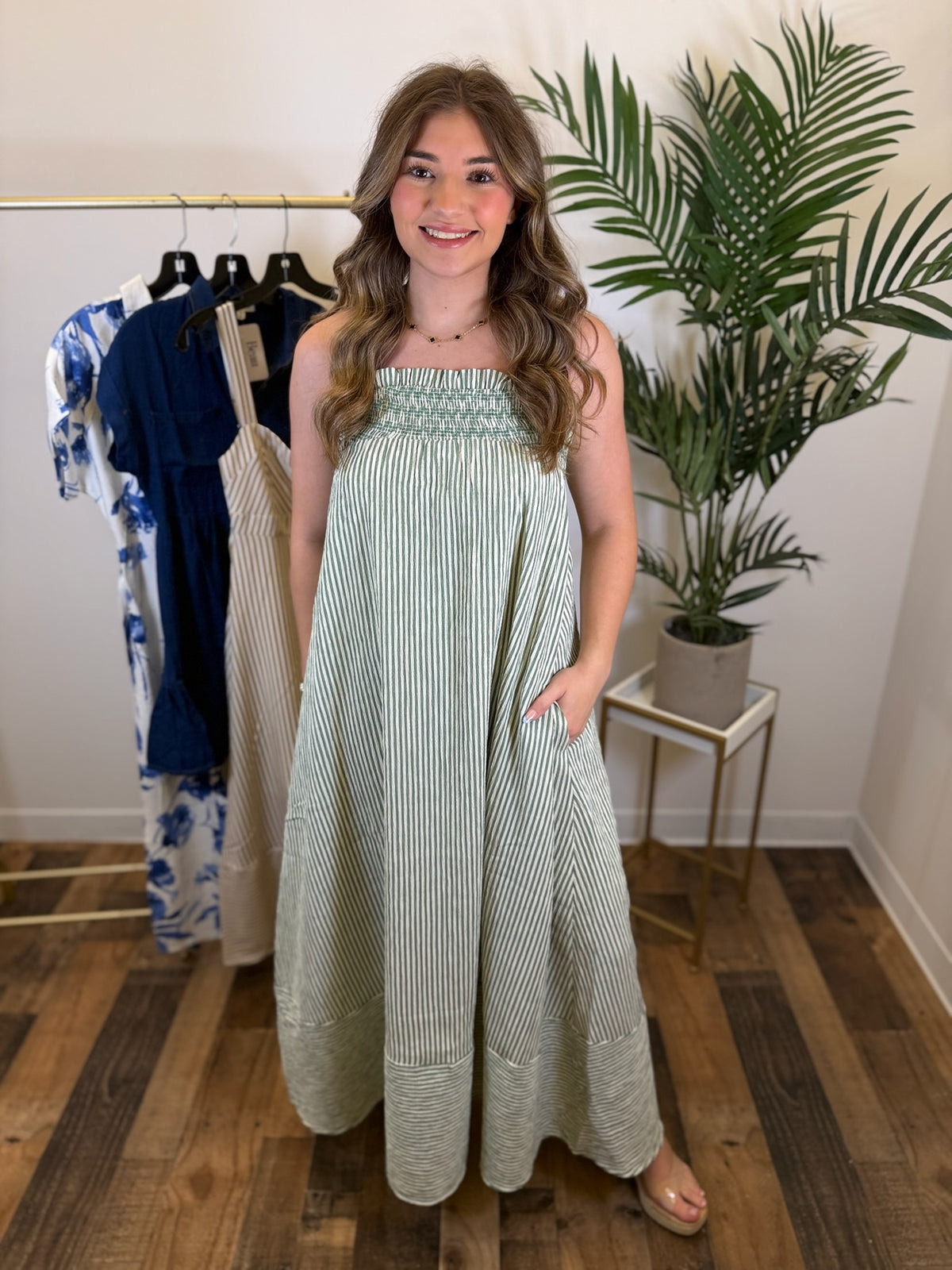 Woman wearing a sleeveless green striped maxi dress with smocked neckline standing in a room with a clothing rack and plant.