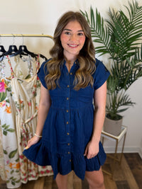 Woman wearing a blue dress standing in a clothing store with floral dresses and plants in the background.