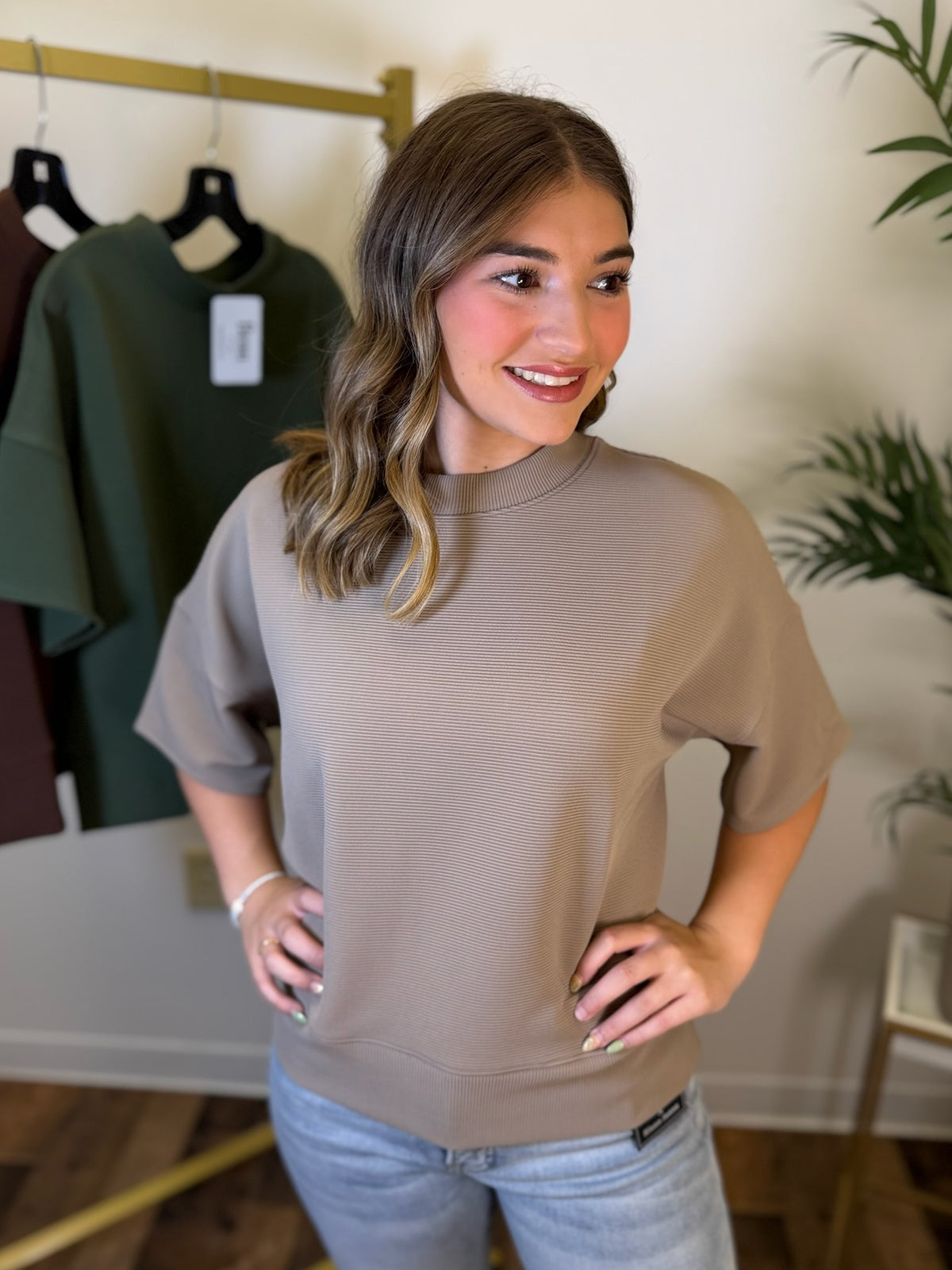 A woman wearing a taupe short-sleeve crewneck sweatshirt and light blue jeans, standing in a boutique setting with a clothing rack and plants in the background.