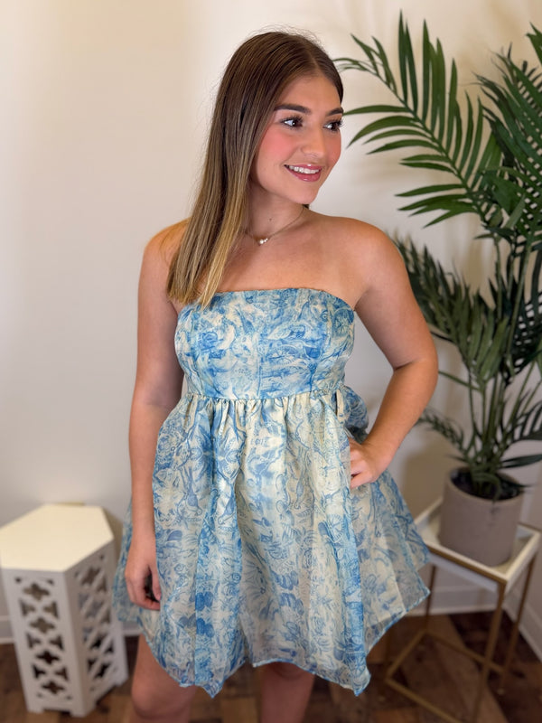 A woman modeling a strapless light blue and ivory toile-patterned mini dress in an indoor setting with a potted plant in the background.