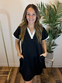 Woman wearing a short sleeve black shift dress with white trim standing indoors next to a plant with her hands in pockets.