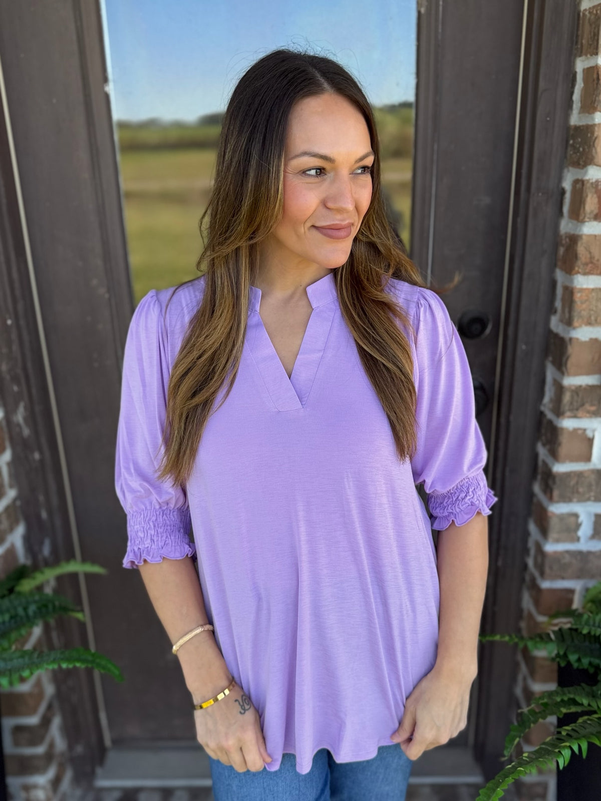 Woman wearing a lavender blouse with short puff sleeves, smocked detailing, and a split V-neckline.