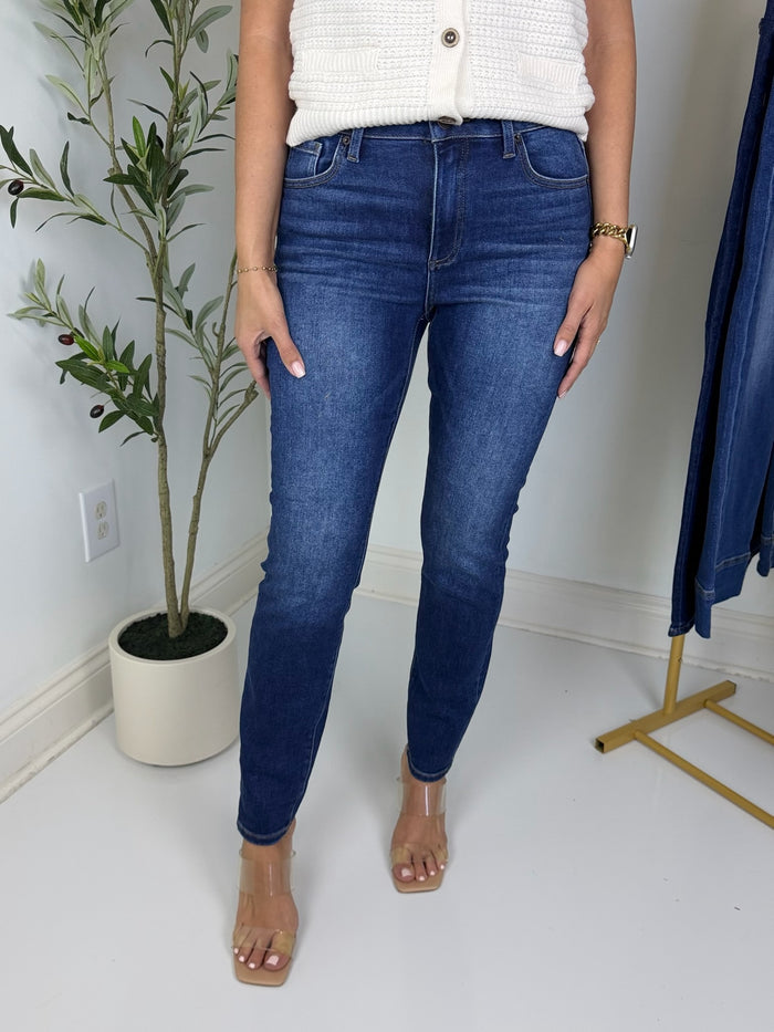woman wearing dark wash skinny jeans in white room with clothes rack and plant in background.